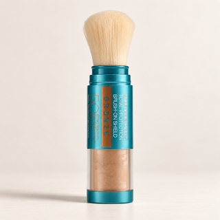 A single makeup brush with a teal metallic handle, a clear lower section showing bronze powder, and cream-colored bristles, standing upright on a light beige background.