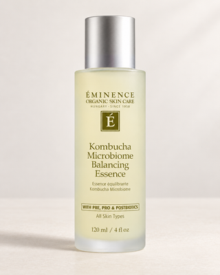A bottle of Éminence Organic Skin Care Kombucha Microbiome Balancing Essence with a frosted glass body, pale yellow liquid, and a metallic silver cap, set against a clean, light beige background.