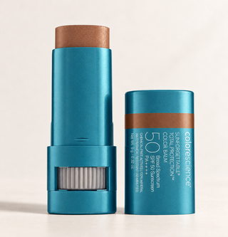 Two teal-blue sunscreen color balm sticks from colorscience stand on a light beige studio background. One stick is open, showing the copper-brown balm bullet and a white filter mesh window near the base.