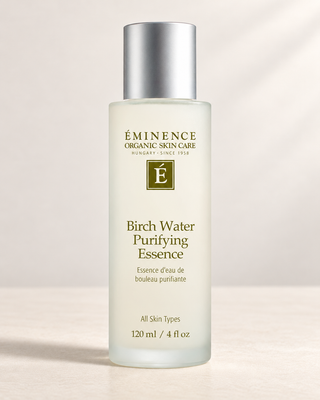 A bottle of Éminence Organic Skin Care Birch Water Purifying Essence, featuring a frosted glass bottle with a metallic silver cap, set against a soft beige background with subtle diagonal shadows.