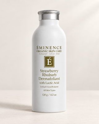 A white cylindrical bottle of Éminence Organic Skin Care Strawberry Rhubarb Dermafoliant with Lactic Acid, featuring a silver screw cap and gold lettering, set against a warm beige background with soft shadows.