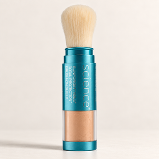 A single makeup brush with a teal metal handle and a clear lower section showing a warm beige powder inside, photographed on a clean off-white background. The handle has visible text including 'science' and 'total protection brush shield.