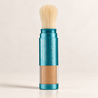 A single makeup stick brush with a teal metallic body, clear lower section showing tan powder, and a soft cream-colored brush bristle on top, photographed on a clean light beige background.